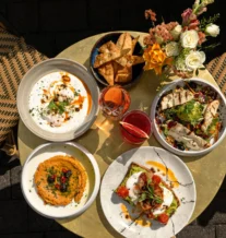 A sunny outdoor table with appetisers, drinks, and vibrant flowers at a welcoming café.