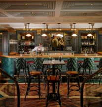 A cosy hotel bar with a bartender serving drinks to guests seated at the counter.