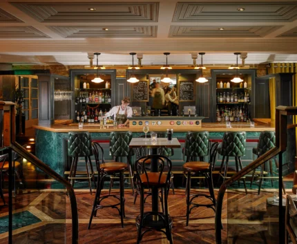 A cosy hotel bar with a bartender serving drinks to guests seated at the counter.