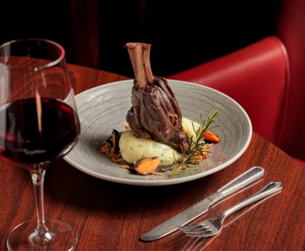 Elegant lamb shank dish with wine in a warm, upscale restaurant setting.