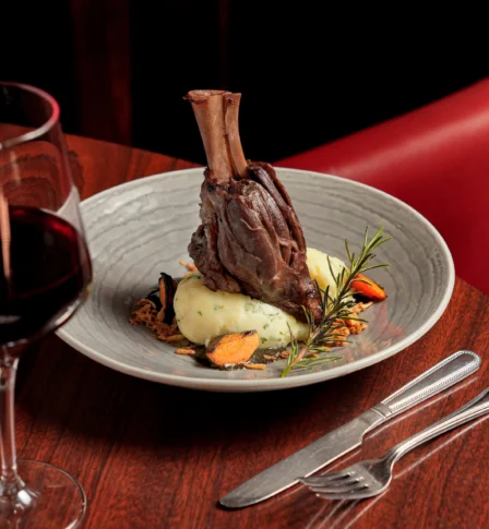 Elegant lamb shank dish with wine in a warm, upscale restaurant setting.