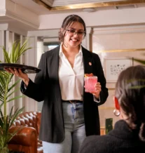 Friendly server offers pink cocktail to guests in cosy hotel lounge.
