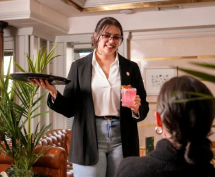 Friendly server offers pink cocktail to guests in cosy hotel lounge.