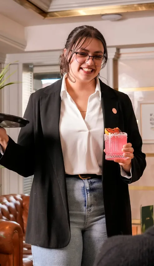 Friendly server offers pink cocktail to guests in cosy hotel lounge.
