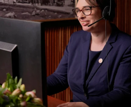 Woman at desk smiling while assisting guests via headset in a hotel office.