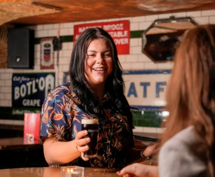 A woman in a cosy pub enjoys a Guinness with a friend, surrounded by vintage signs.