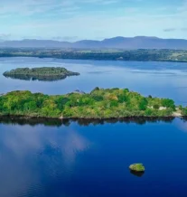 Scenic lake view with lush green island and distant mountains under blue sky.