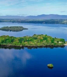 Scenic lake view with lush green island and distant mountains under blue sky.