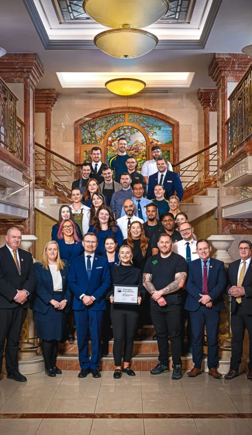 A welcoming hotel team stands on an elegant staircase, holding a framed award.