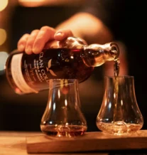 Warm-lit bar with whisky being poured into two glasses on a wooden board.