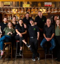 Smiling staff pose warmly at a cosy pub counter with wooden decor and hanging glasses.