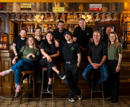 Smiling staff pose warmly at a cosy pub counter with wooden decor and hanging glasses.
