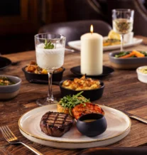 Cosy dining table with steak, sides, candle, and drinks.