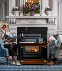 A couple relax by a cosy hotel fireplace, enjoying books and drinks in an elegant lounge.