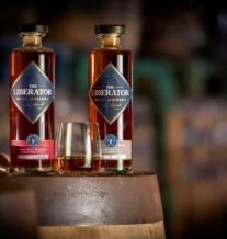 Two bottles of The Liberator Irish Whiskey on a barrel with a glass, in a cosy setting.