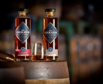 Two bottles of The Liberator Irish Whiskey on a barrel with a glass, in a cosy setting.