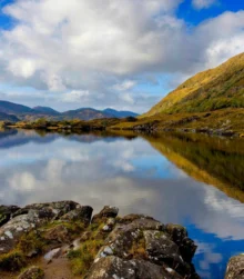 A person stands by a tranquil lake, surrounded by lush hills and a clear blue sky.