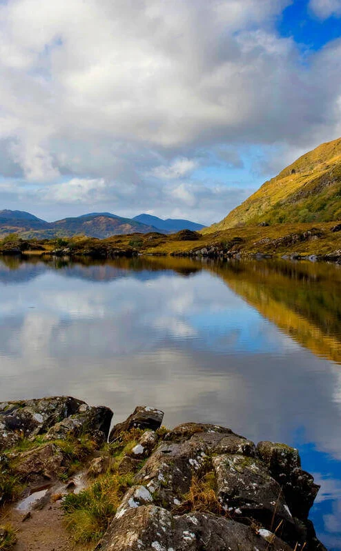 A person stands by a tranquil lake, surrounded by lush hills and a clear blue sky.