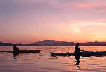 A couple kayaking on a tranquil lake at sunset with mountain views.