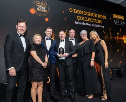 Smiling hotel team family in evening wear holding a Gold Medal Hotel Awards trophy on stage, backdrop reads "O'Donoghue Ring Collection" and "Ireland's Best Hotel Group