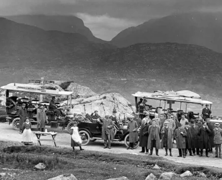 Family group and couples beside vintage motor coaches on a misty mountain road, picnic table and rocky landscape visible, signage on coach reads "LOCHALSH & WEST HIGHLAND TOUR