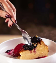 A hand reaches to enjoy berry-topped cheesecake on a plate in a cosy dining setting.