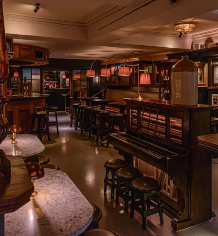 Warm, cosy pub interior with a piano, marble-topped tables, and ambient lighting.
