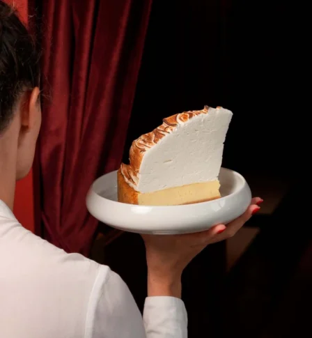 Person holding a plate with a large slice of lemon meringue pie by a window with red curtains.