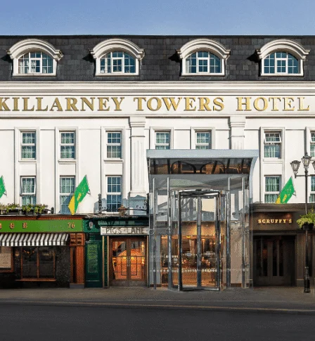 Family arriving at Killarney Towers Hotel entrance, warmly lit façade with flags and street café (80 characters)