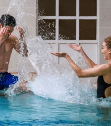 Family splashing joyfully in an indoor pool at a hotel, playful couple creating big water spray
