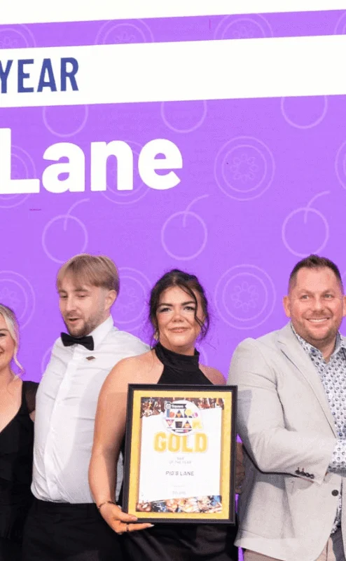 Smiling group of family and friends on stage accepting a "Bar of the Year" award for Pig's Lane, purple backdrop